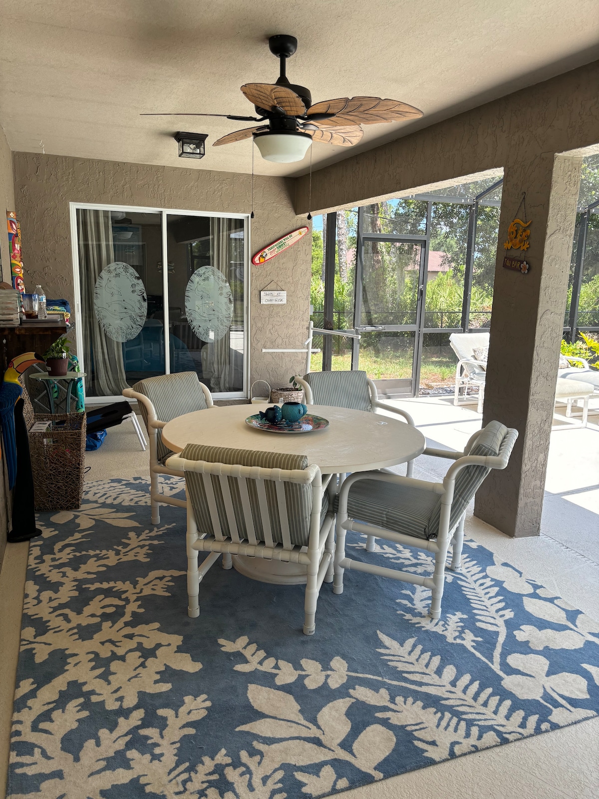 A screened-in outdoor seating area is showcased, featuring a round table surrounded by four white chairs. A patterned blue rug covers the floor, and a ceiling fan adds comfort. Large sliding doors provide views of the garden and pool area, enhancing the space's openness.