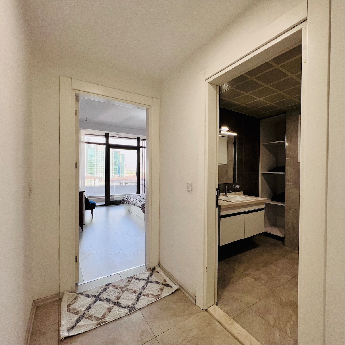 A spacious hallway is visible, leading to a living area illuminated by natural light. An open doorway on one side reveals a well-equipped bathroom with modern fixtures. The walls are painted in neutral tones, enhancing the overall bright atmosphere of the space.