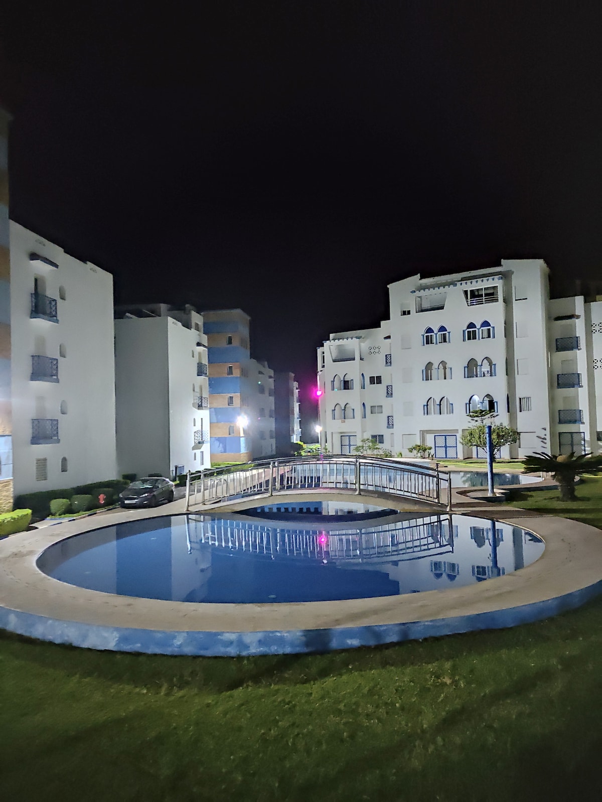 The nighttime view captures a serene courtyard featuring a circular pool, reflecting the surrounding buildings. Soft lighting enhances the garden areas and provides a tranquil ambiance, with the residential structures creating a sense of enclosure.