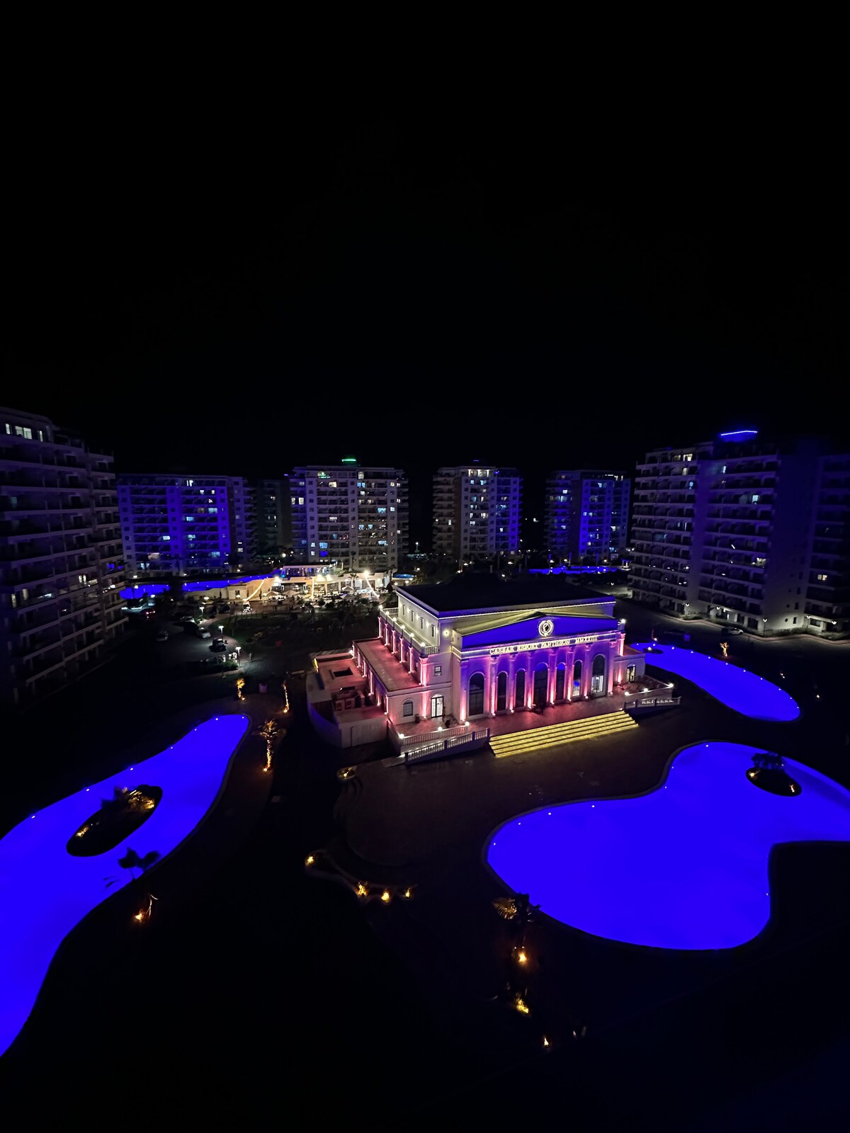 An elegant building stands illuminated at night, showcasing a blend of purple and blue lighting. Surrounding areas are designed with curved water features, reflecting the vibrant colors. Apartment-style buildings are visible in the background, contributing to a lively atmosphere.