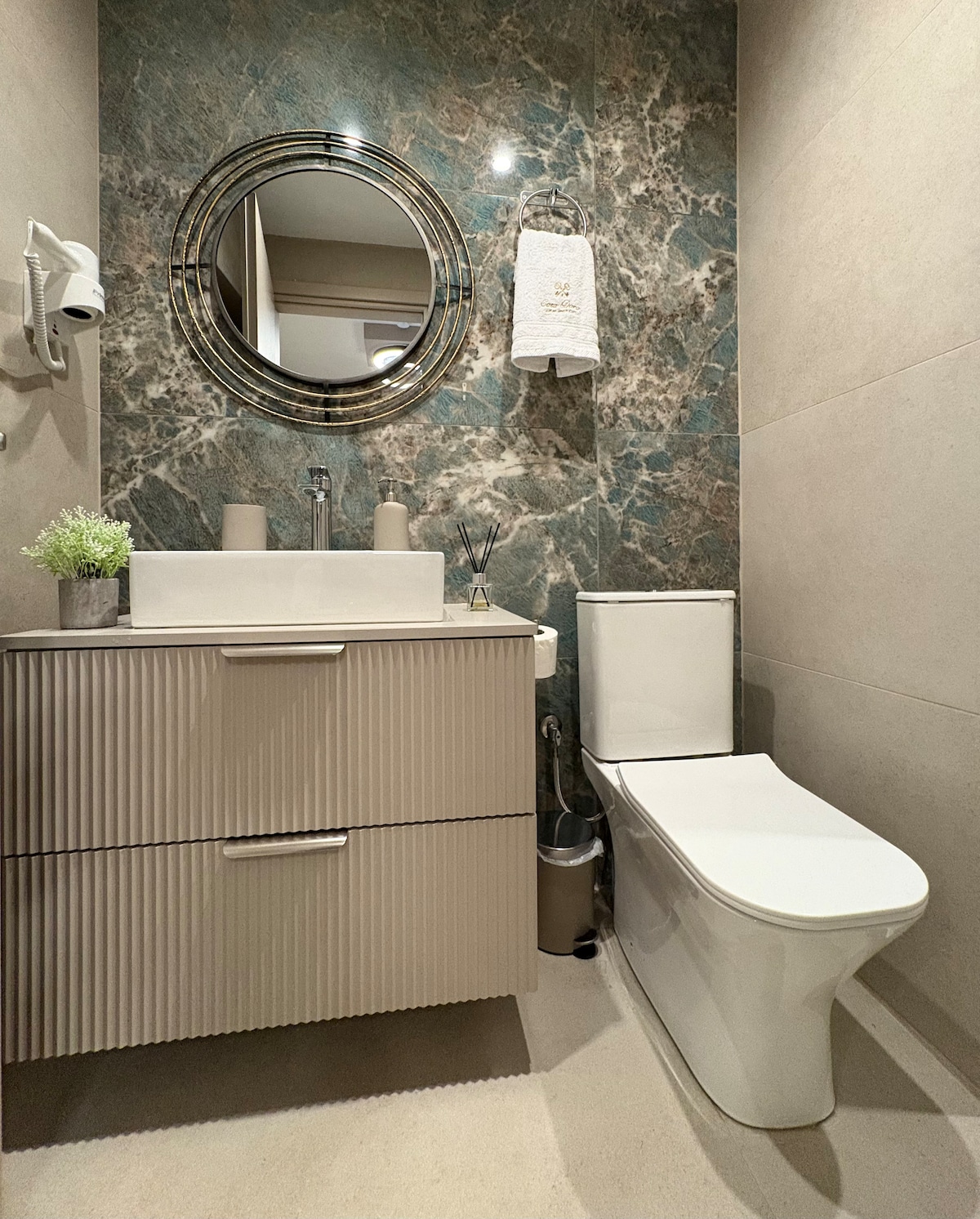 The bathroom features a modern vanity with vertical ridging, providing a textured element against the marble-like wall. A round mirror is mounted above the sink. A soft plant accentuates the counter, while a compact toilet is positioned to the side, completing the clean design.