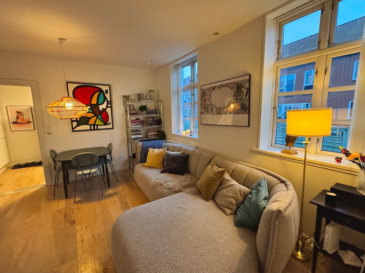 A living area features a light-colored sectional sofa adorned with colorful cushions, providing ample seating. A small dining table with gray chairs sits nearby. Natural light filters through large windows, illuminating wall art and a bookshelf filled with various items.