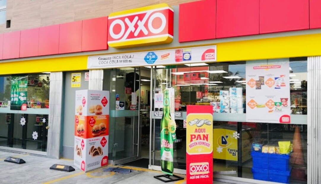A convenience store entrance is featured, displaying a colorful store awning. The entrance is flanked by promotional displays and glass doors, showcasing the items available for purchase. Various products, including beverages and snacks, can be seen on the exterior stands.