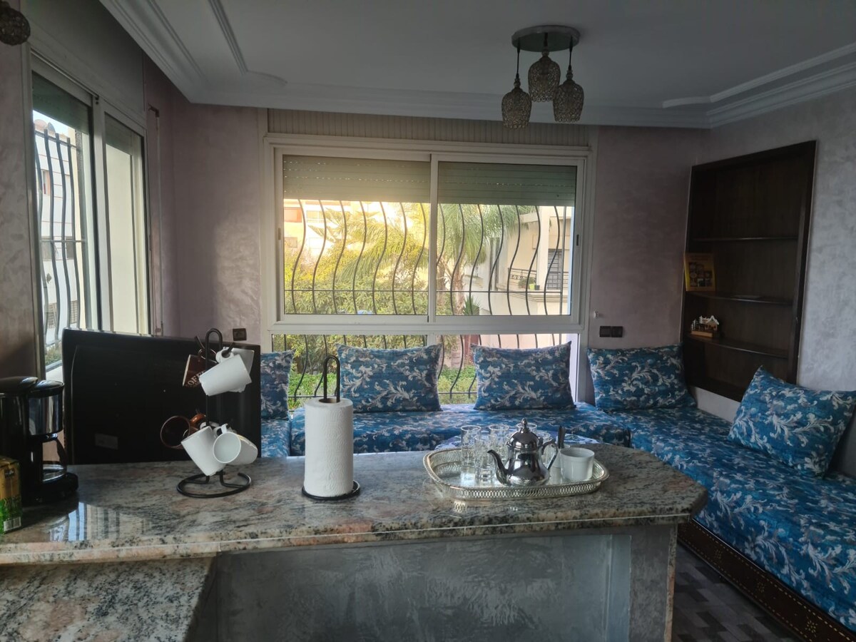 A modern kitchen area features a granite countertop with a coffee maker and decorative tray. A cozy seating area with a blue-patterned sofa is set near large windows, allowing natural light to fill the space. Stylish light fixtures hang from the ceiling, enhancing the ambiance.