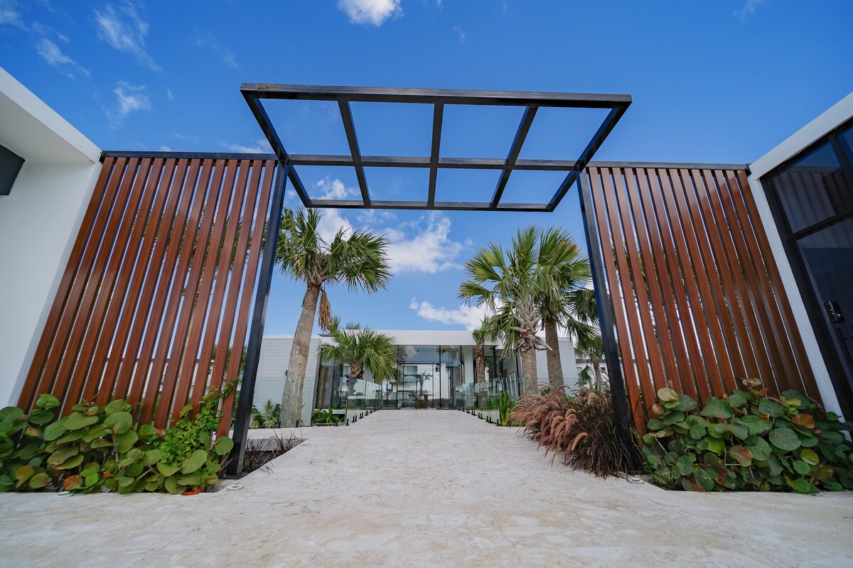 A modern entrance features a stylish framework supported by wooden slats and a pathway lined with tropical foliage. Palm trees provide a lush backdrop against a clear blue sky, inviting guests into the serene atmosphere of the property.