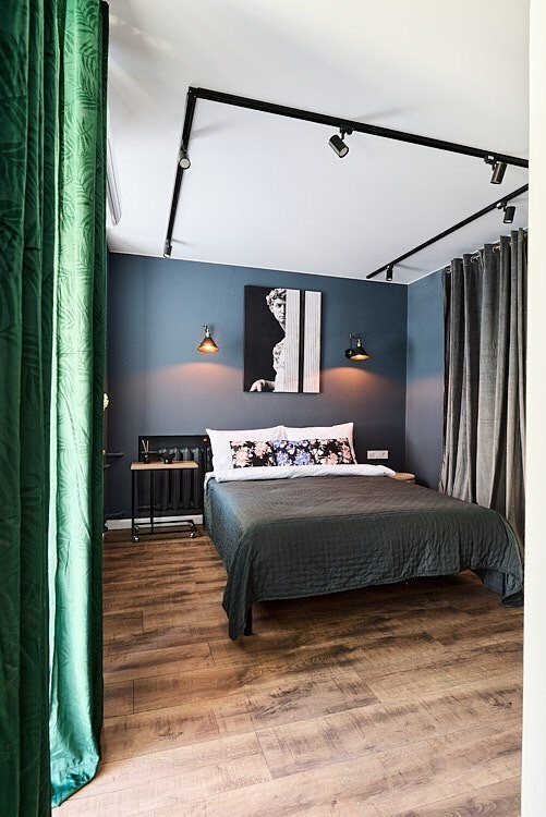 A compact sleeping area features a cozy bed with a soft, dark coverlet. Wall-mounted lights provide focused illumination above the bed. Artistic wall décor and rich curtains enhance the room's character, while warm wood flooring adds a touch of comfort.