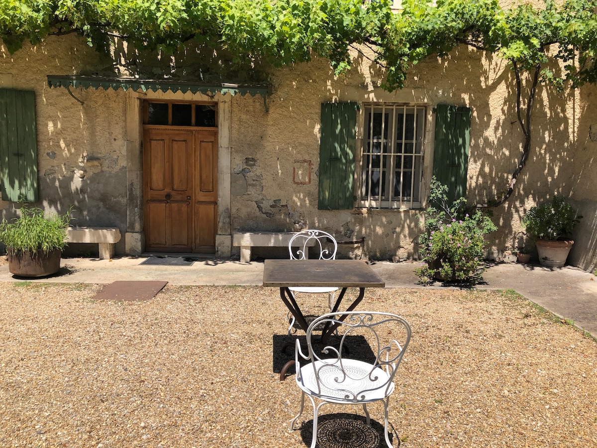 A quaint outdoor space features a small table and two chairs set on gravel, surrounded by lush greenery. The rustic stone wall of the gîte is adorned with green shutters and a door, creating a calm and inviting atmosphere for al fresco dining.