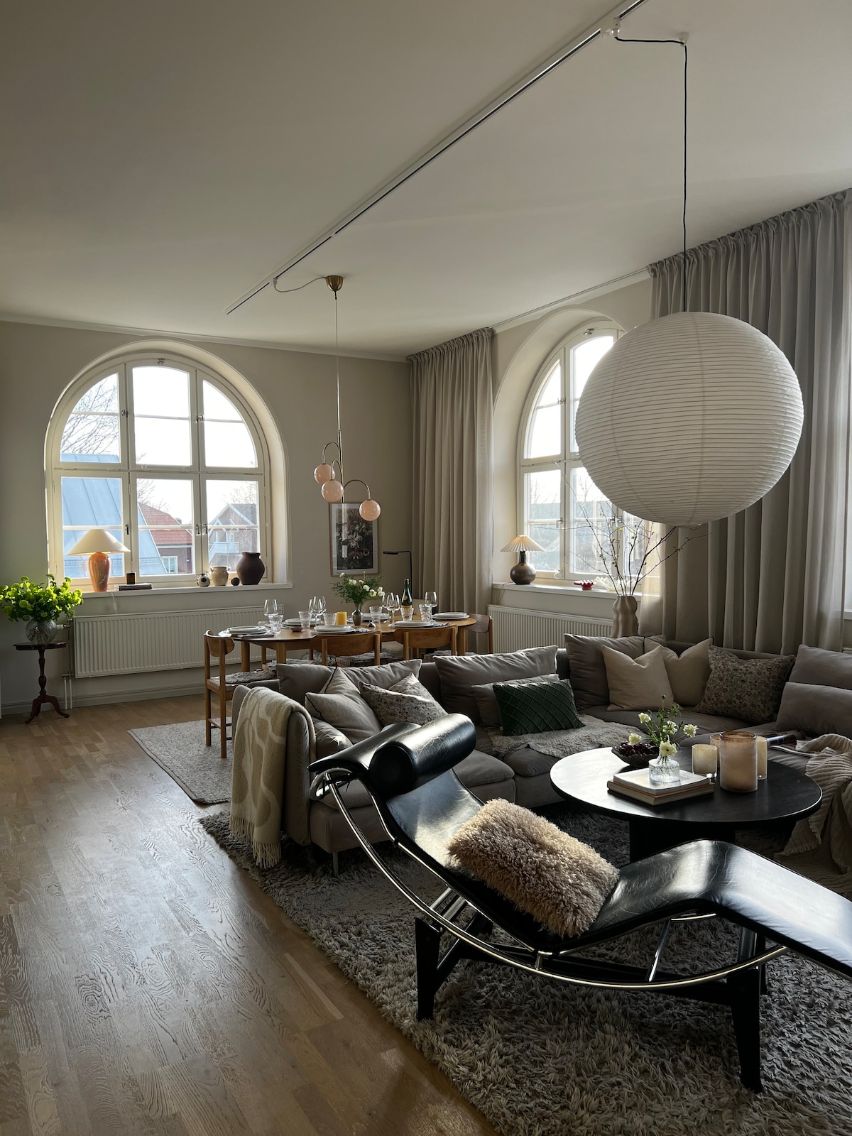 A spacious living area features a large sectional sofa adorned with various textured cushions. A circular table set for dining is visible nearby, alongside large windows that allow natural light to fill the room. Soft curtains and decorative lighting create a calming ambiance.