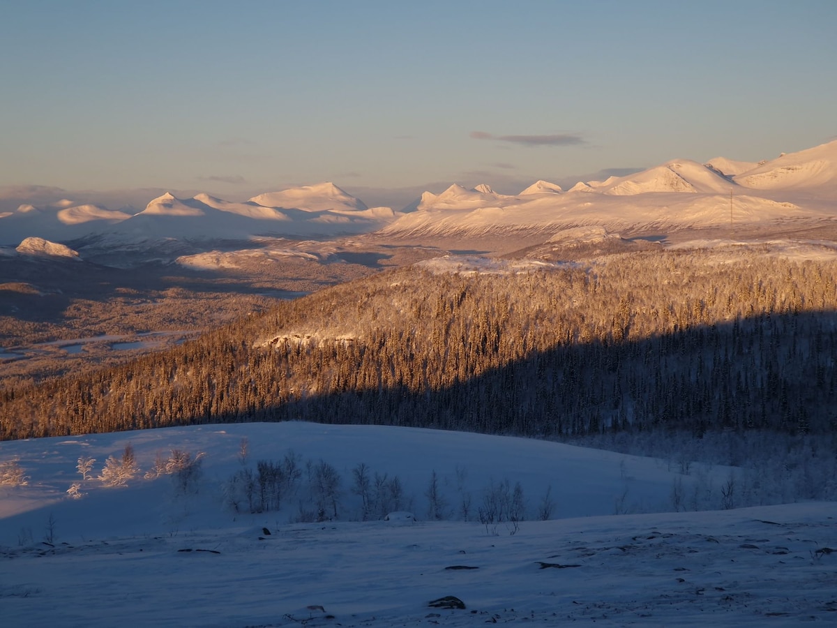 A scenic view of snow-covered mountains under a clear sky. The landscape features rolling hills and a mix of dark shadows and bright snow, highlighting the natural contours. Sunlight casts a soft glow over the peaks, creating a serene winter atmosphere.