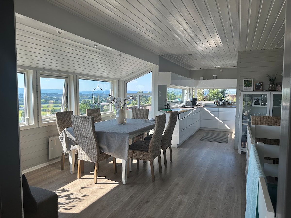 A bright dining area is visible, featuring a large table surrounded by six chairs. Large windows provide scenic views of the surrounding landscape, allowing natural light to fill the space. A white kitchen is adjacent, with a minimalist design and ample cabinetry.