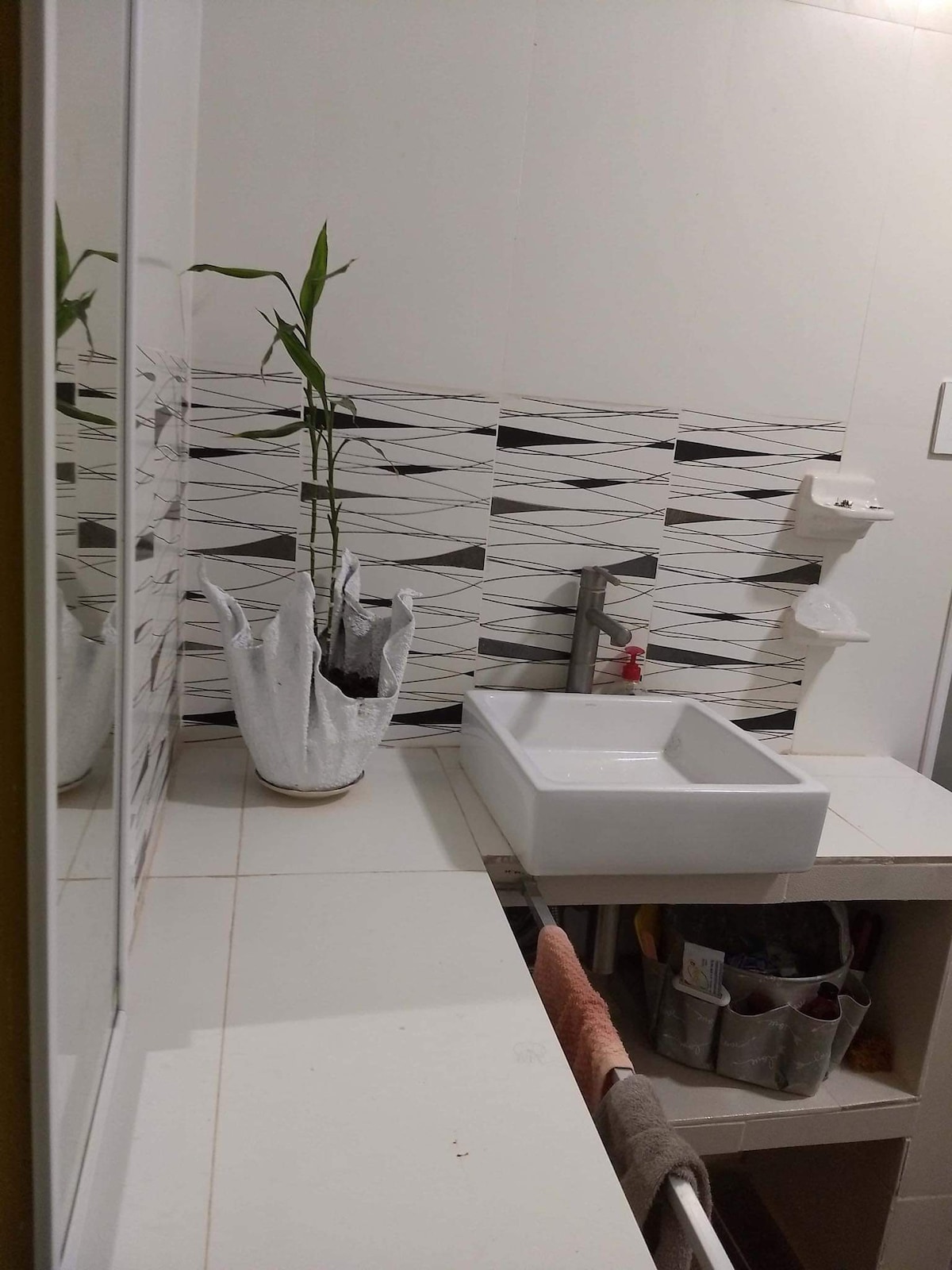 A modern bathroom features a sleek white sink atop a minimalist countertop, complemented by patterned wall tiles. A small plant sits in a white pot, adding a touch of greenery. Shelving is provided for toiletries, while the overall layout emphasizes clean lines and a tidy atmosphere.