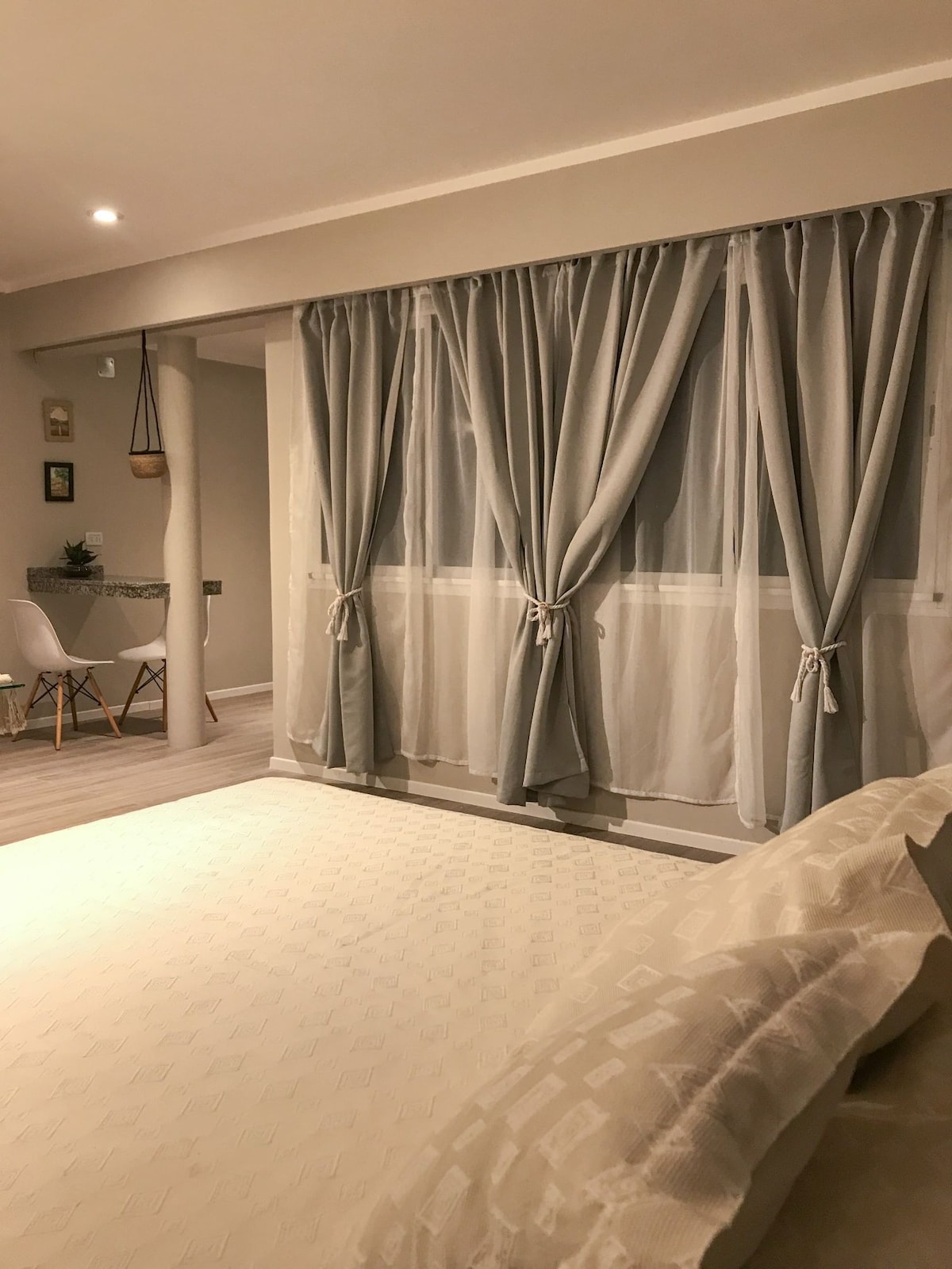 A spacious room is highlighted by large, soft gray curtains that elegantly frame a window. A small dining area with a round table and two chairs is visible in the corner, complementing the light, neutral color palette of the space.