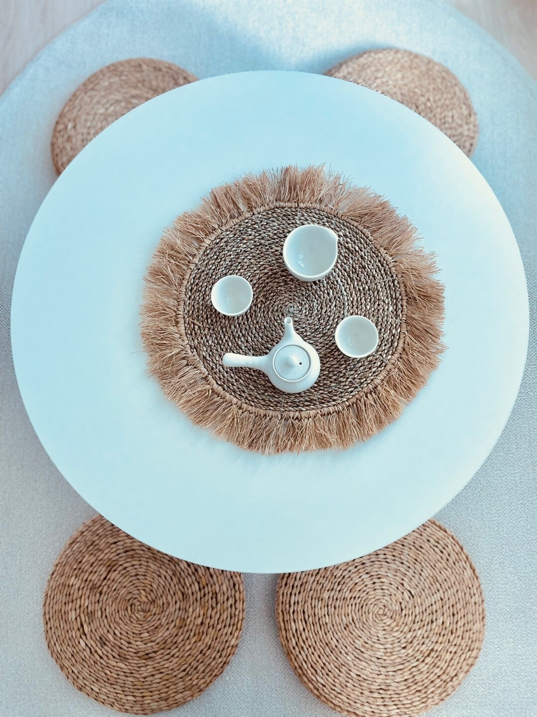 A round dining table is set with a light blue surface. A woven centerpiece surrounds a teapot and four small cups, all arranged on a natural woven placemat. Circular woven mats are placed under each chair, creating a cohesive design.