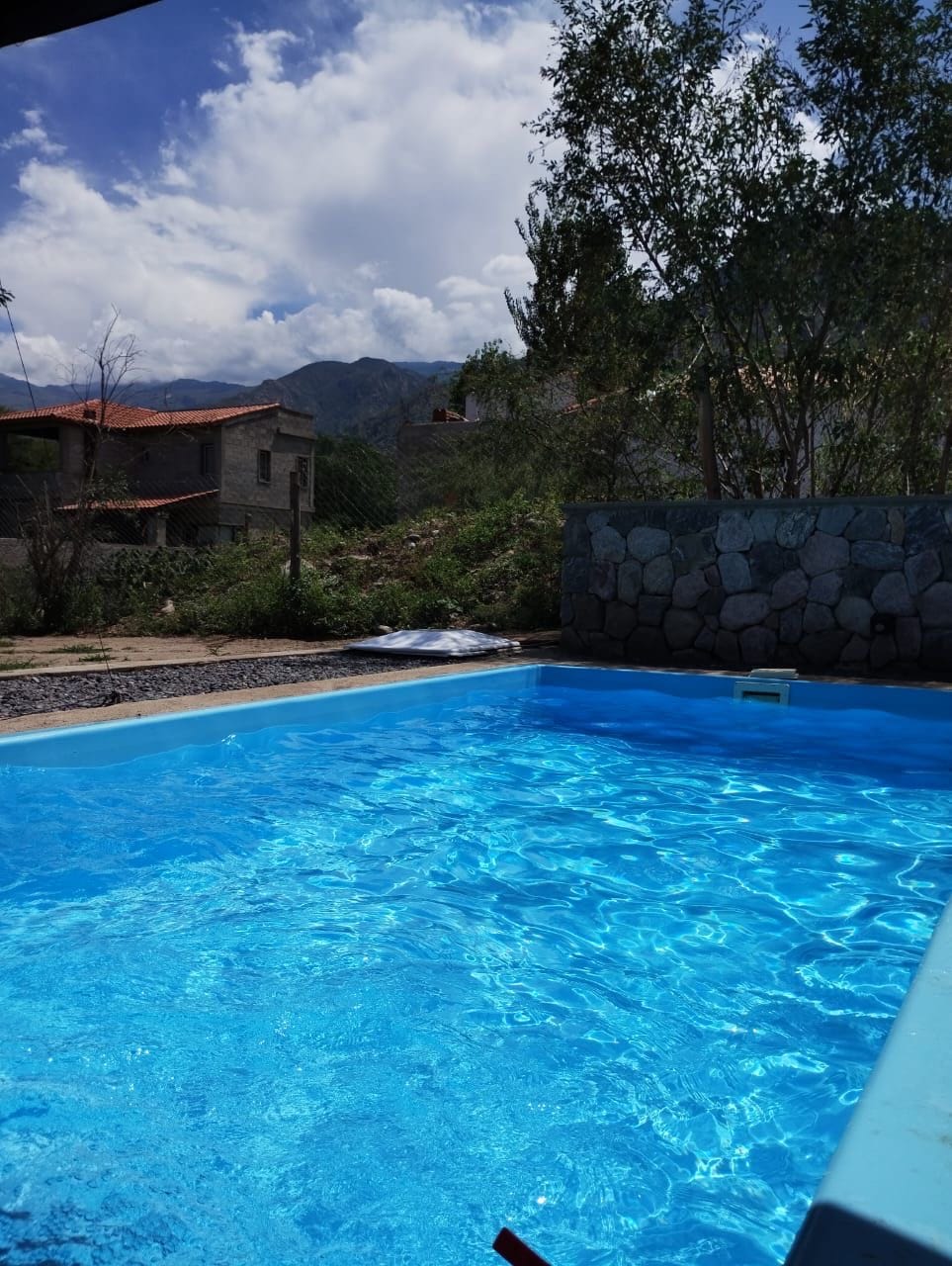 A clear blue swimming pool is pictured, reflecting the sky above. Surrounding greenery and stone walls provide a natural backdrop. In the distance, traditional buildings are visible, set against a mountainous landscape.