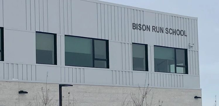 The exterior of Bison Run School is visible, featuring a modern design with large windows that allow natural light to enter. The building's name is prominently displayed on the upper façade, reflecting a clean and minimalist architectural style.