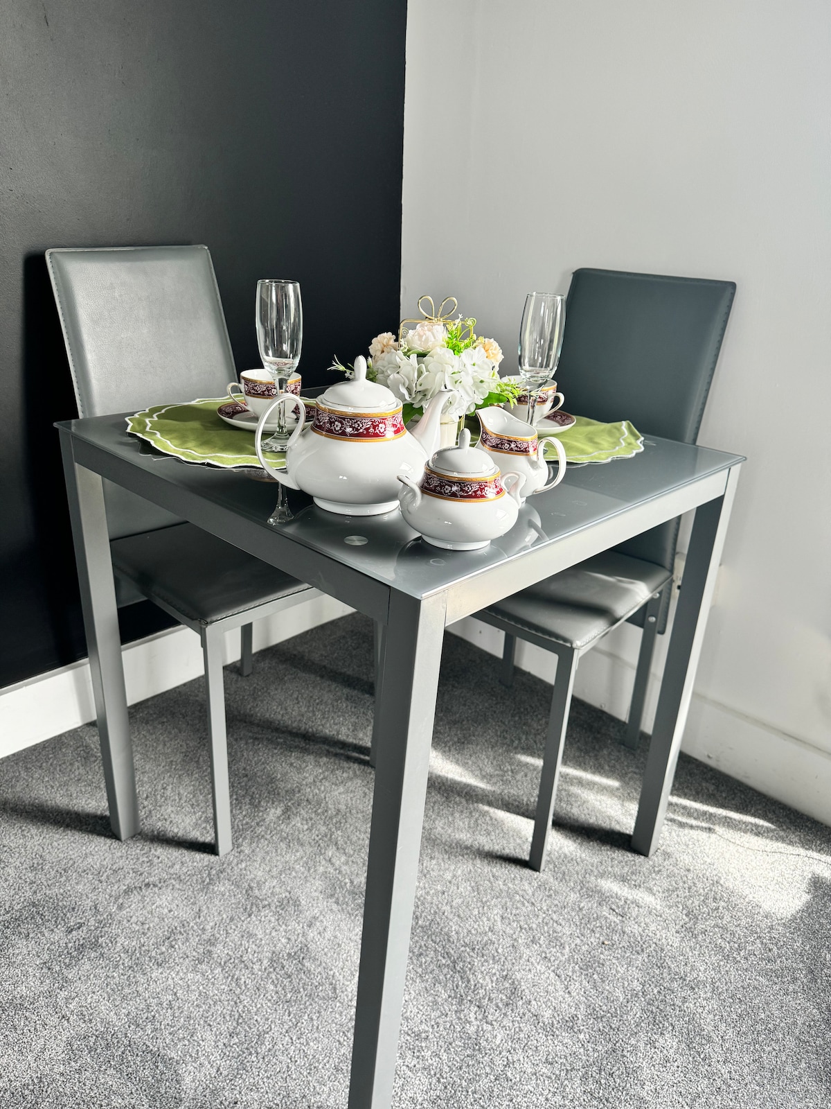 A compact dining table is set with fine china and decorative dishware. Two elegant chairs are positioned alongside the table. A floral centerpiece adds a touch of freshness, while glasses and teapots are arranged neatly to create an inviting dining experience.
