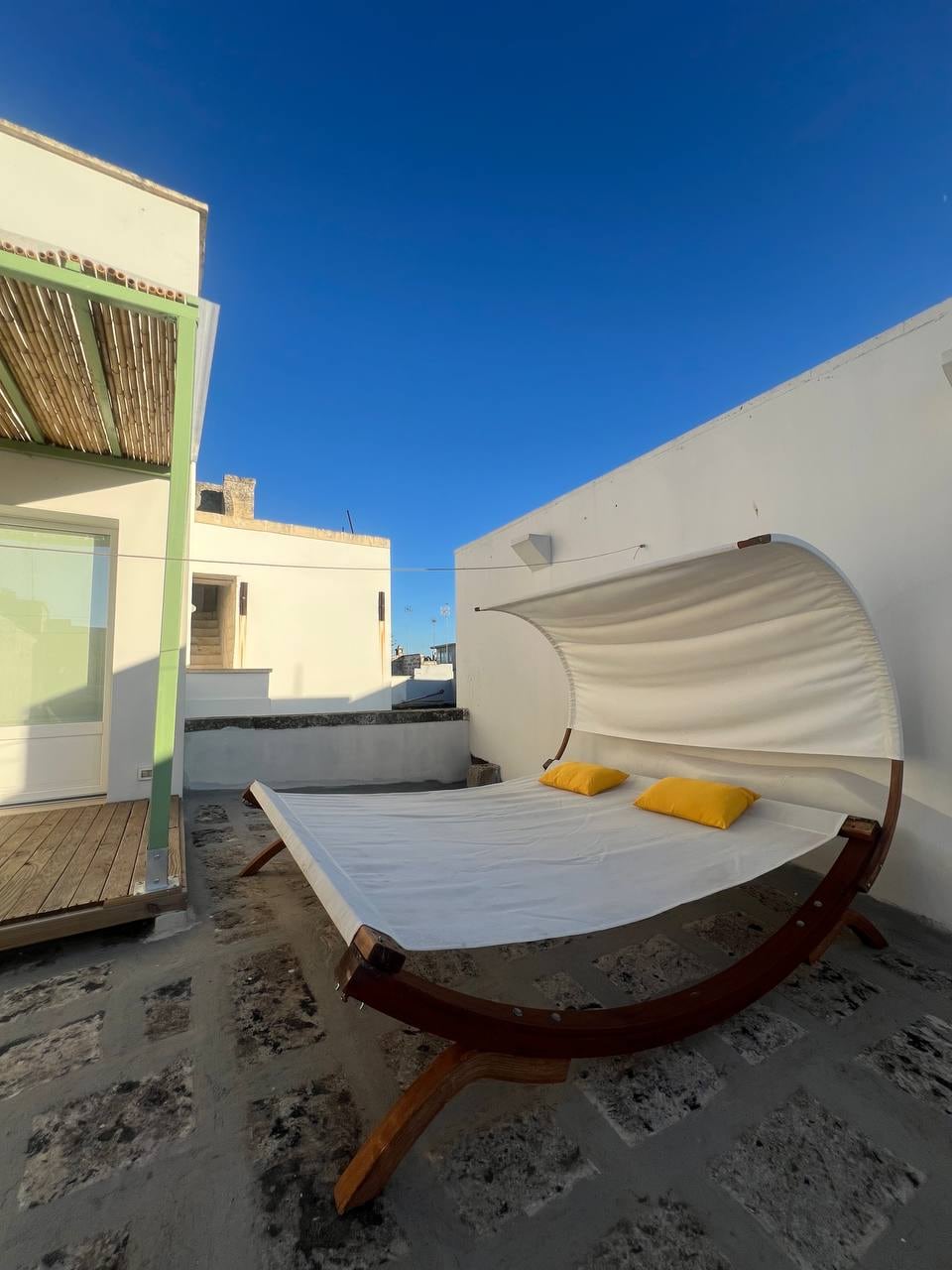 A sleek outdoor lounging area features a curved wooden bed with two yellow cushions. The unit is positioned on a stone surface, with a clear blue sky visible above and a light structure nearby, providing an inviting space for relaxation.