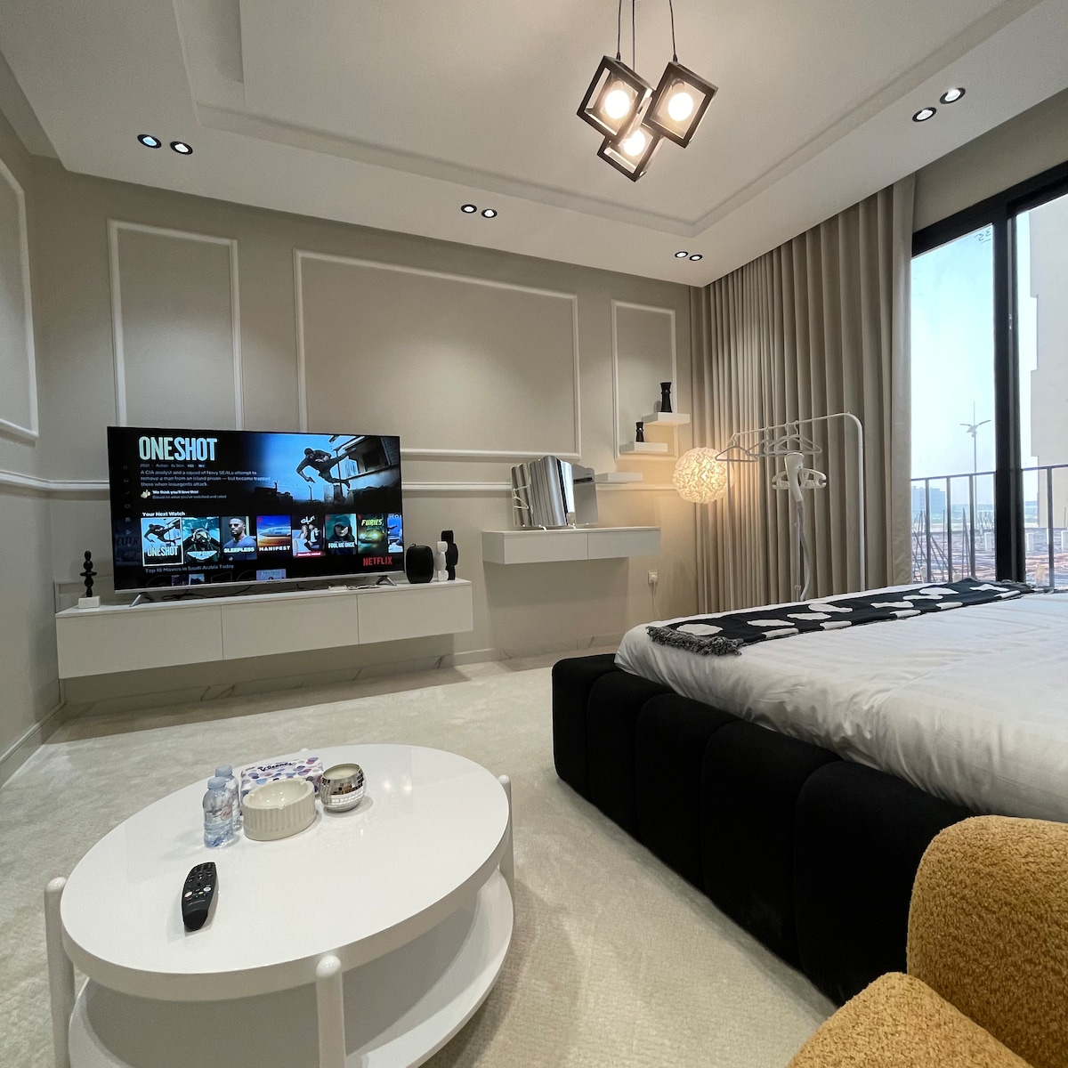 A modern studio features a large television displaying content options. A spacious bed is positioned beside a round coffee table with decorative items. Natural light floods through a floor-to-ceiling window, with a stylish light fixture hanging from the ceiling.
