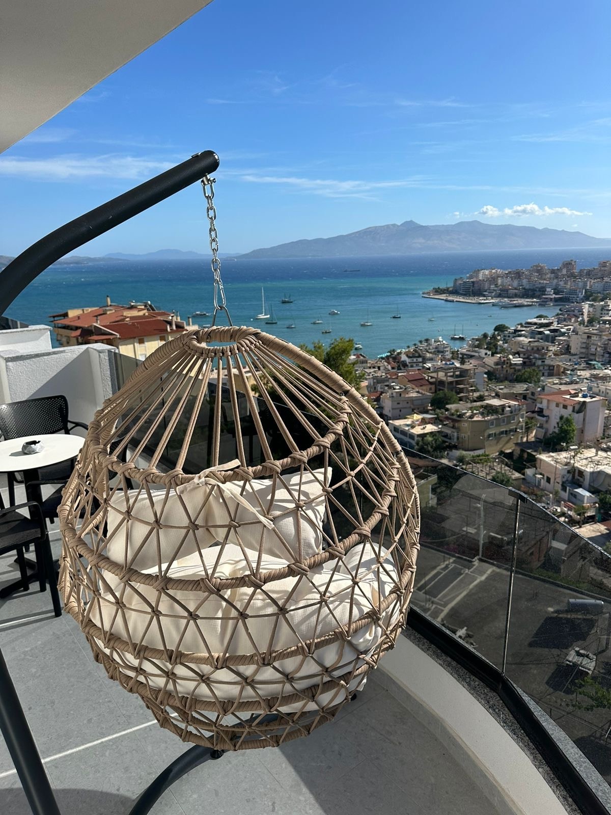 A hanging chair with soft cushions is positioned on a balcony, offering stunning views of a coastal landscape. The turquoise water meets the horizon adorned with boats. A small bistro table and chairs complement the outdoor space, providing a relaxing atmosphere.