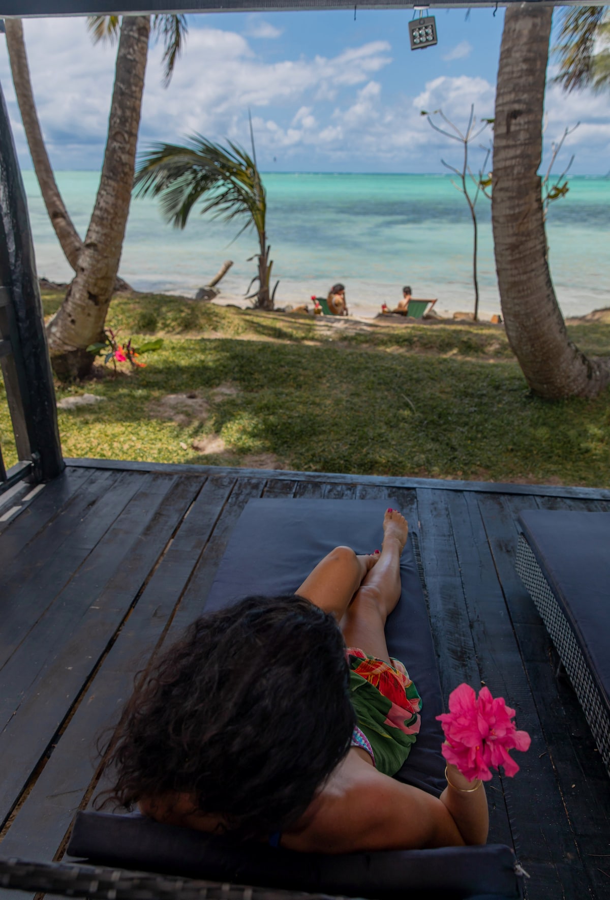 A serene view from a shaded deck, showcasing a person reclining on a lounge chair, holding a pink flower. In the background, palm trees frame a sandy beach and turquoise waters, where a few people are enjoying the calm environment.