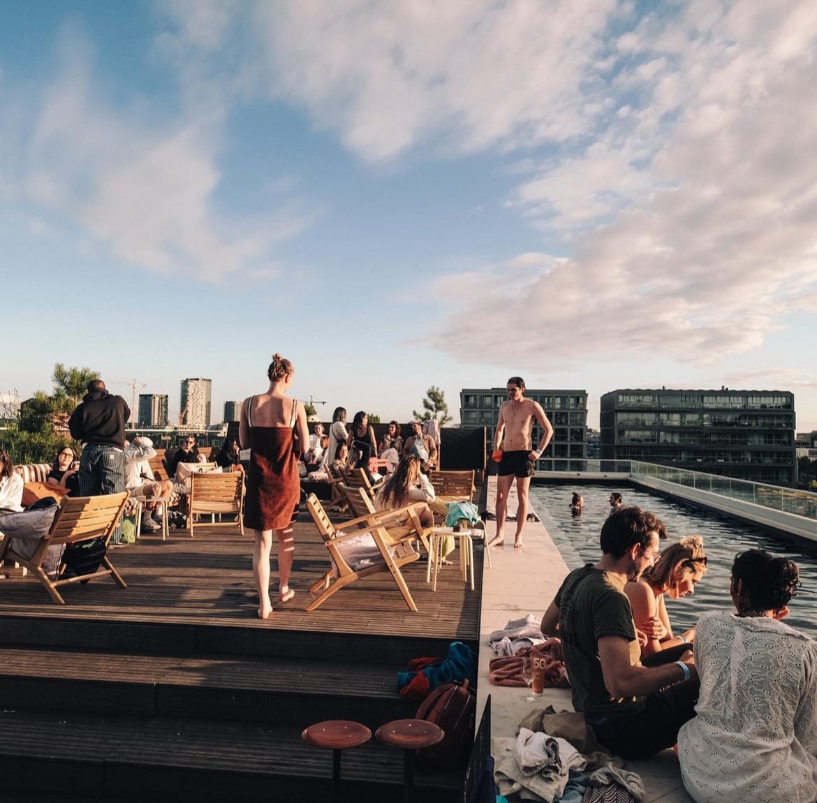 A vibrant rooftop scene captures a diverse group of people enjoying a sunny day. Comfortable lounge chairs and a swimming pool are visible, along with a clear sky scattered with clouds. People are seen socializing by the poolside and relaxing on the deck.