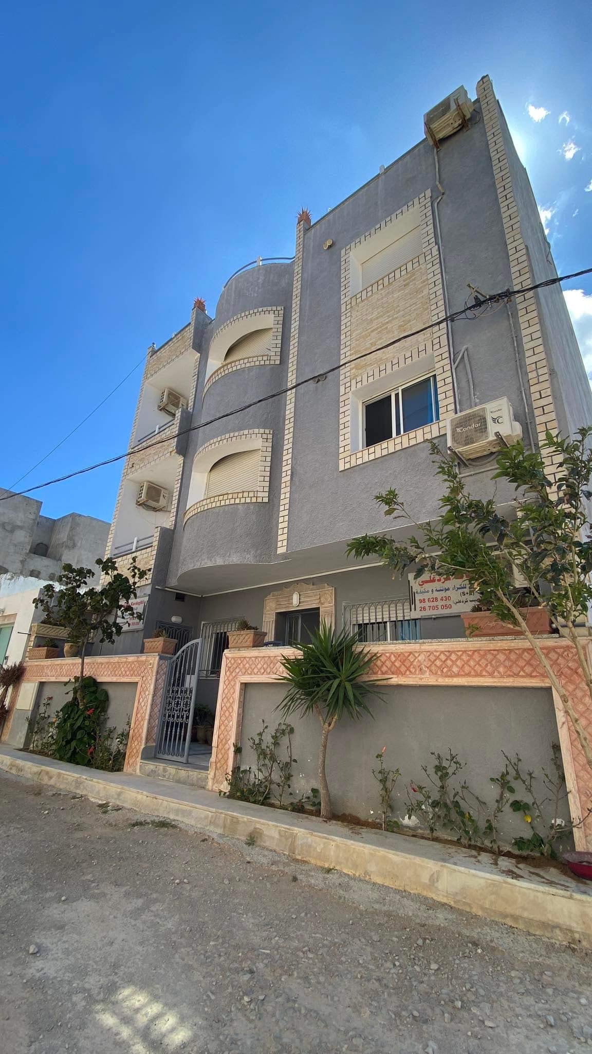 The exterior of a three-story gray building is presented, featuring decorative brickwork and multiple air conditioning units. A small garden with shrubs and plants surrounds the entrance, adding a touch of greenery. Bright blue skies add to the setting's inviting appearance.