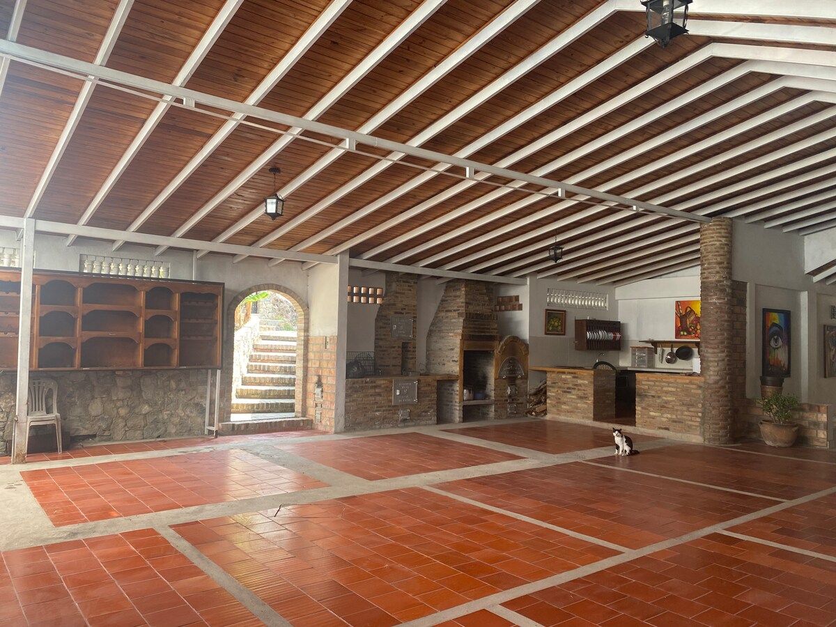 An expansive outdoor space features a tiled floor with a warm terracotta hue, complemented by a wooden ceiling with exposed beams. Stone walls and shelves create an inviting area, while a staircase leads to a different section, enhancing the openness of the environment.