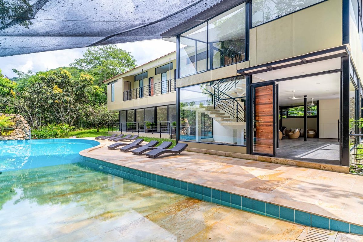 A modern two-story house is depicted next to a clear swimming pool. Lounge chairs are arranged along the poolside. Large glass windows and doors promote natural light and views of the green surroundings, enhancing the connection between indoor and outdoor spaces.