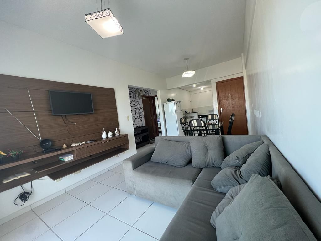 A modern living area features a neutral-colored sectional sofa with several cushions. A wall-mounted television is positioned above a sleek wooden shelf, and the space is illuminated by overhead lighting. An open kitchen area is visible in the background, showcasing dining furniture.