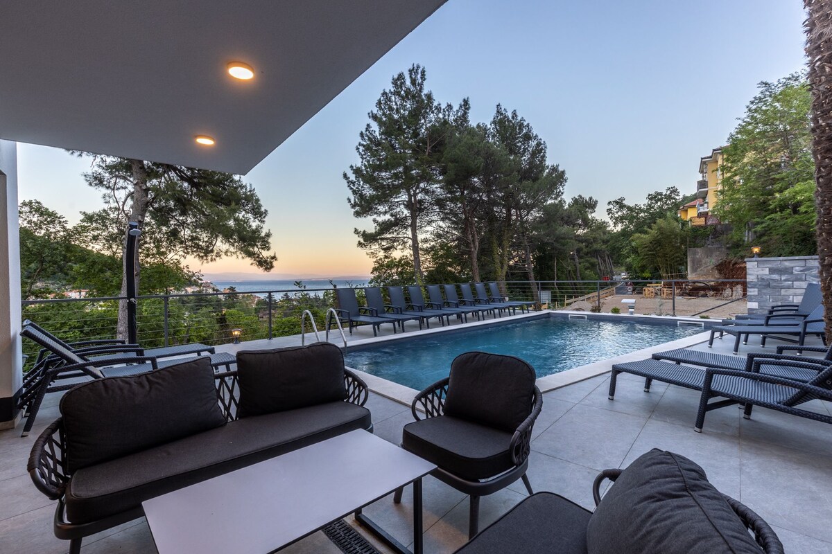 A poolside area is furnished with multiple lounge chairs and dark outdoor seating. The swimming pool reflects the evening sky, surrounded by lush greenery and residential buildings in the background. Soft lighting enhances the serene atmosphere as dusk approaches.