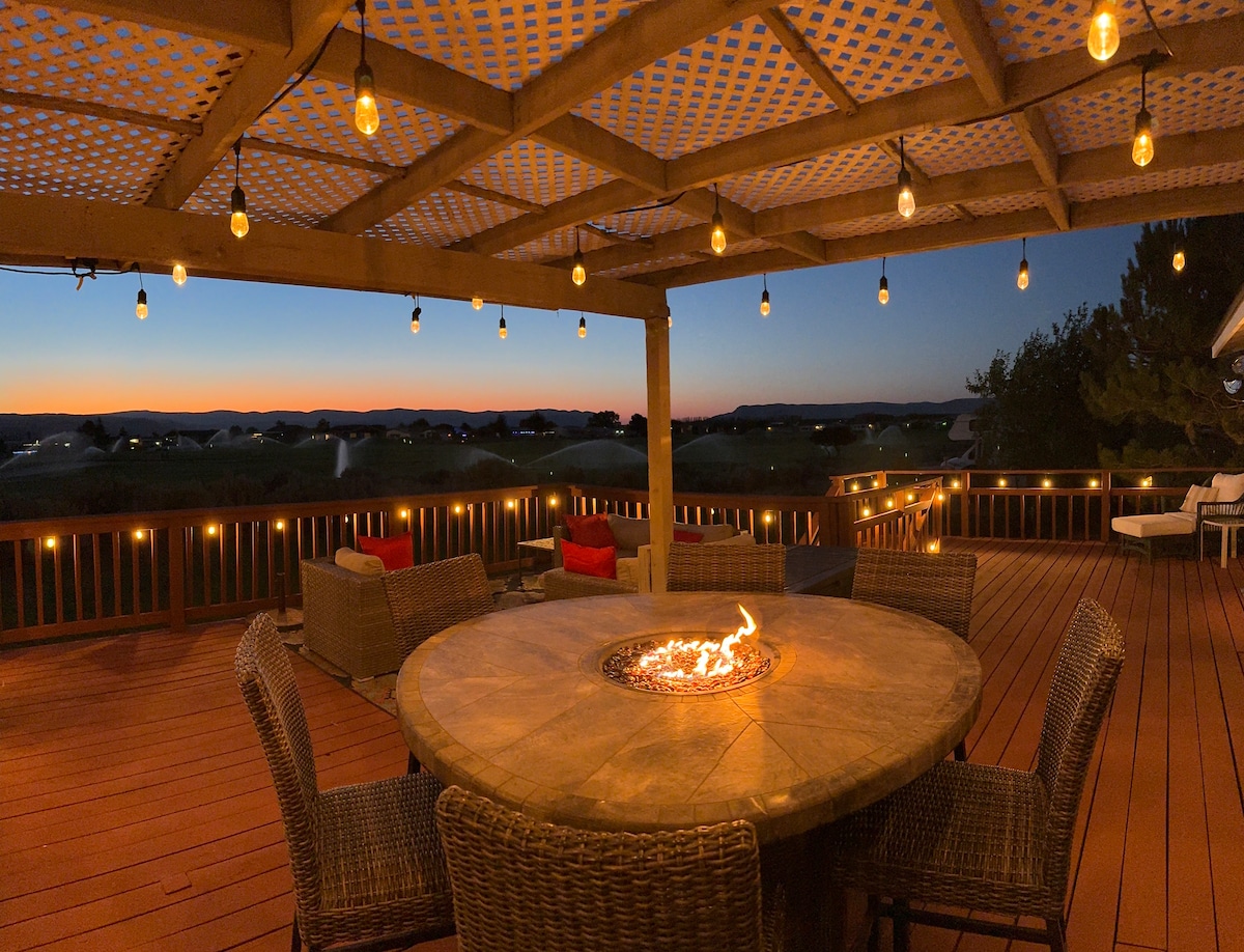 An outdoor deck area features a round stone fire pit at its center, surrounded by woven seating. String lights create a warm glow against the evening sky, while the distant silhouettes of rolling hills are visible beyond the railing.