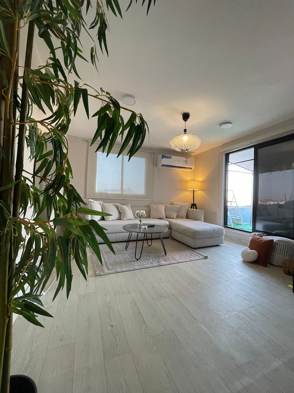 A modern living area features a light-colored sectional sofa and a small black coffee table atop a textured area rug. Natural light fills the space from large windows, and a vibrant indoor plant adds a touch of greenery. A pendant light provides soft illumination.