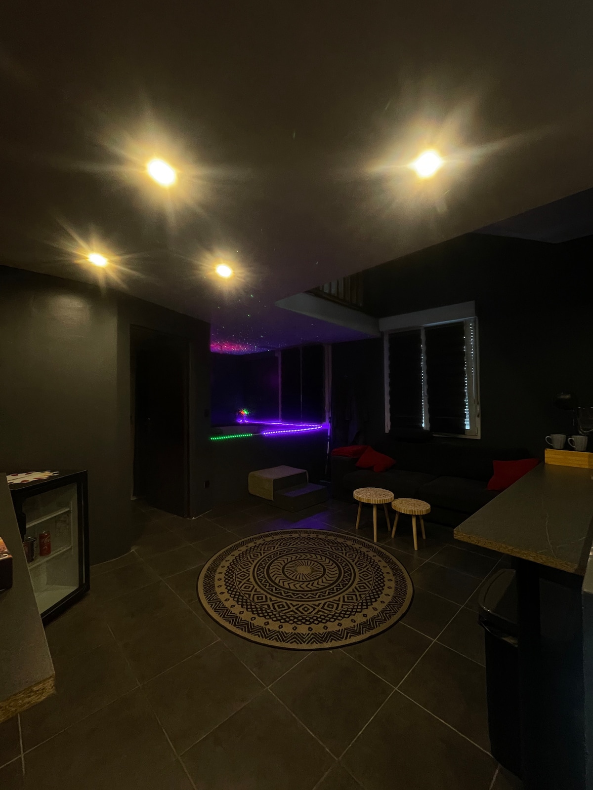 A modern living area features sleek gray walls and ambient lighting from recessed fixtures. A small circular rug lies on the tiled floor, complemented by two minimalist stools next to a low, dark sofa with colorful cushions. Soft hues from a nearby light create a calming atmosphere.