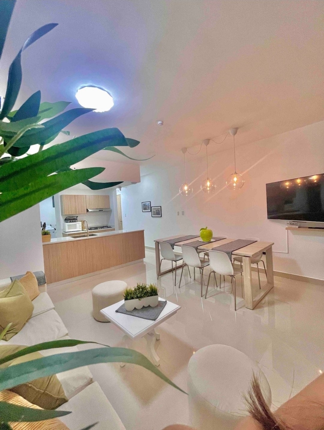 A modern dining area is shown with a sleek table and four chairs, complemented by subtle pendant lighting. A potted plant adds a touch of greenery. In the background, a well-equipped kitchen is partially visible, enhancing the spacious feel of the room.