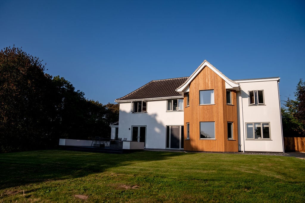 The exterior of Roundwood House is showcased, illustrating a modern two-story structure with a wooden feature wall. Expansive glass windows offer views of the surrounding landscape. A spacious lawn extends in front of the house, providing ample green space for relaxation.