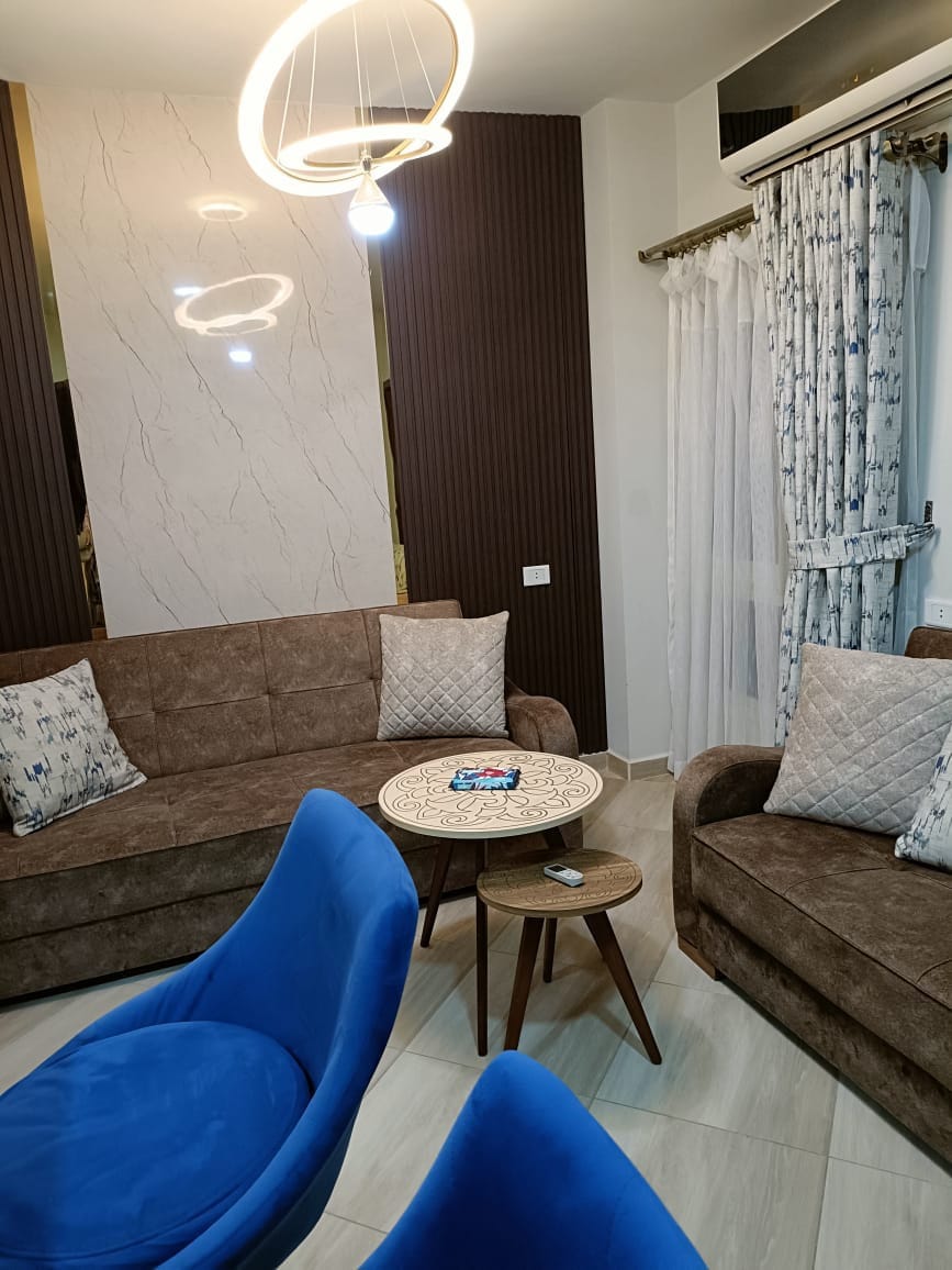 A modern living area is presented with two brown sofas adorned with decorative pillows. A round wooden table sits at the center, complemented by two blue chairs. Soft lighting is provided by overhead fixtures, and sheer drapes frame a window, enhancing the inviting feel of the space.