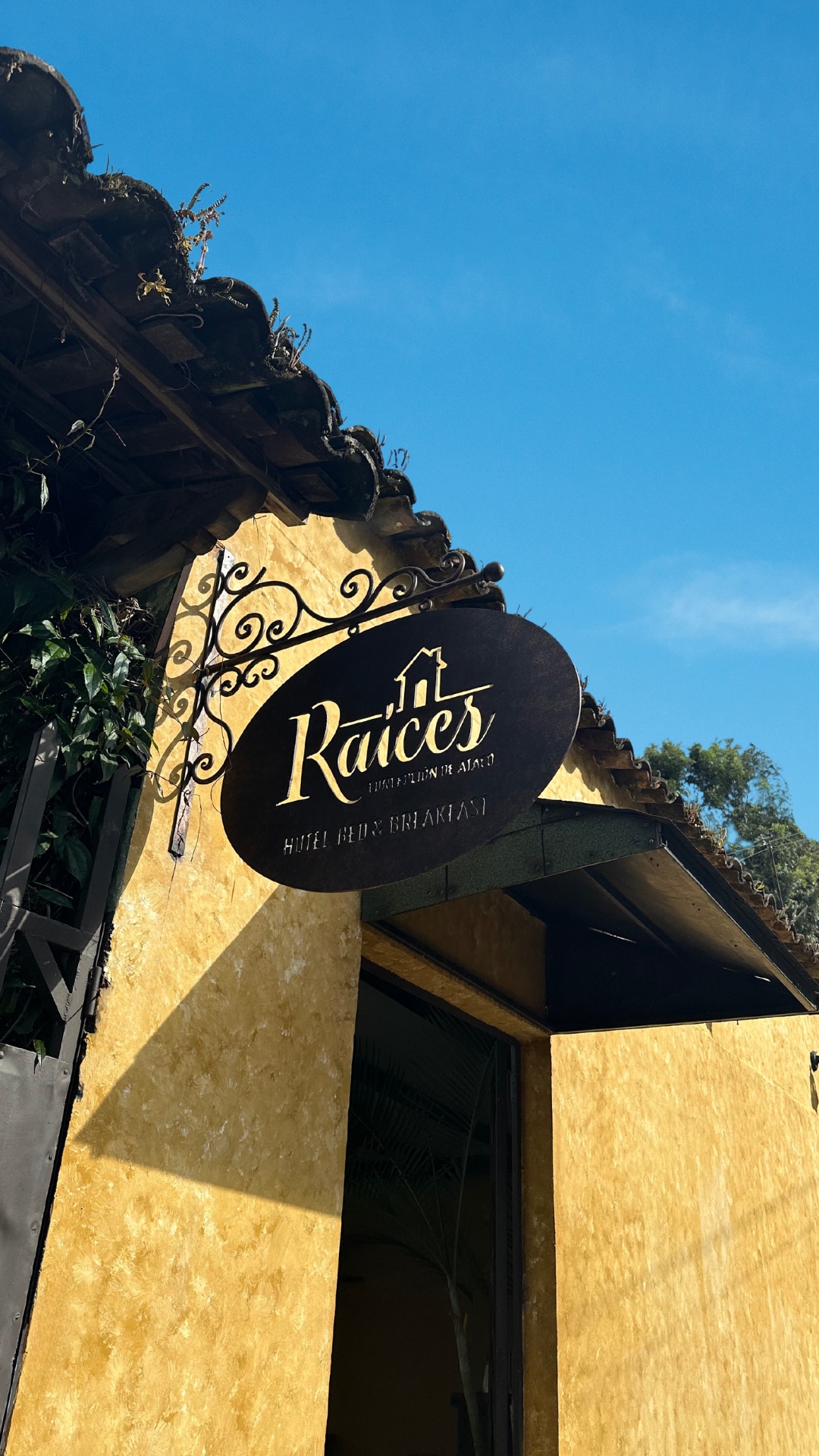 The image displays a sign for Hotel Raíces Ataco, featuring an elegant design with the name prominently displayed. The sign is mounted on a warm yellow wall, surrounded by greenery, set against a clear blue sky.