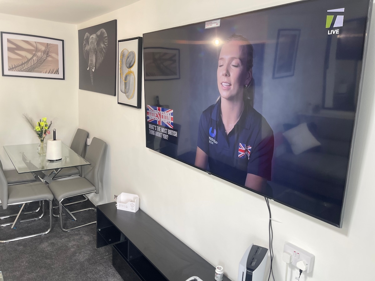 A modern living space is showcased, featuring a wall-mounted television displaying a news broadcast. A glass dining table with six grey chairs is positioned in the foreground, while tasteful wall art, including an elephant print, adds character to the room.