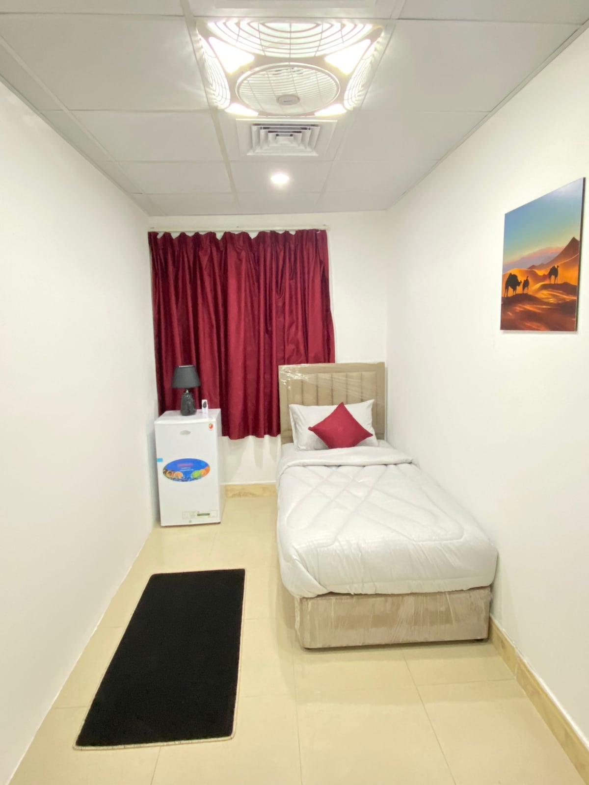 A compact bedroom features a single bed with a light-colored comforter, accompanied by a red accent pillow. A small bedside table is positioned next to the bed with a lamp. A mini-fridge is located nearby, and deep red curtains frame the window, while a piece of wall art adds color.
