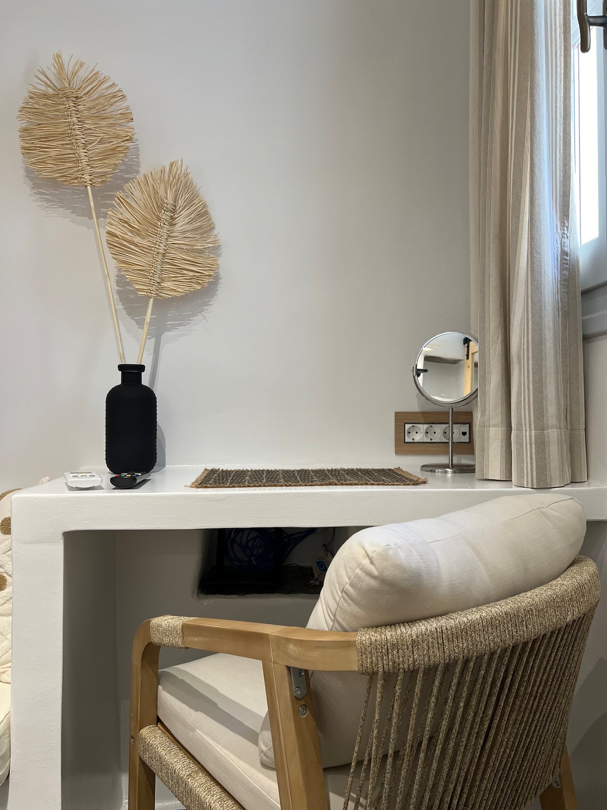 A workstation features a light-colored desk with a textured surface, complemented by a comfortable chair with woven details. Decorative elements include two large, natural-colored wall art pieces and a small black vase. A mirror and electrical outlets are positioned nearby.