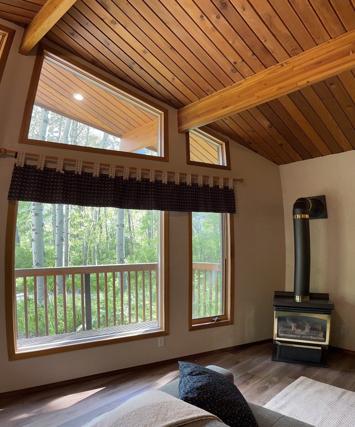 A cozy interior space is highlighted by a high ceiling with wooden beams and large windows that invite natural light. A sleek black wood stove is positioned near the seating area, providing a central focal point while overlooking a tranquil forest view through the balcony.