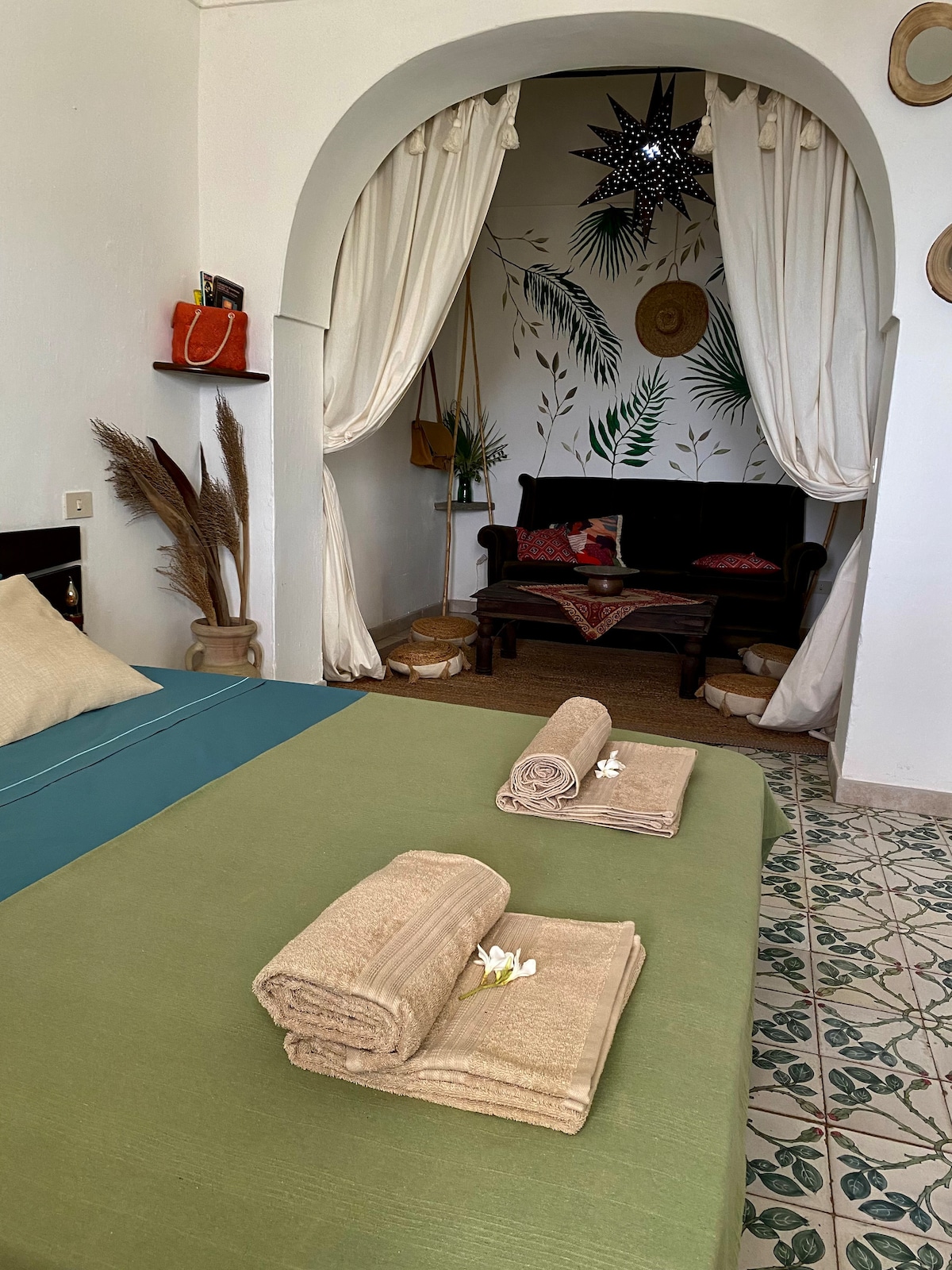 A spacious bedroom features a green bedspread and neatly arranged towels. Soft textiles and natural decor accent the room, complemented by an inviting seating area with a dark sofa. A decorative mural of plants enhances the ambiance, framed by flowing white curtains.