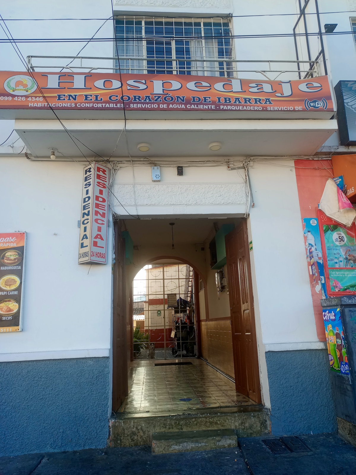 The entrance to a cozy lodging features an archway leading into the interior. Bright signage indicates the establishment's name and services. Flanking the entrance are colorful advertisements and menus from nearby eateries, contributing to a vibrant street scene.
