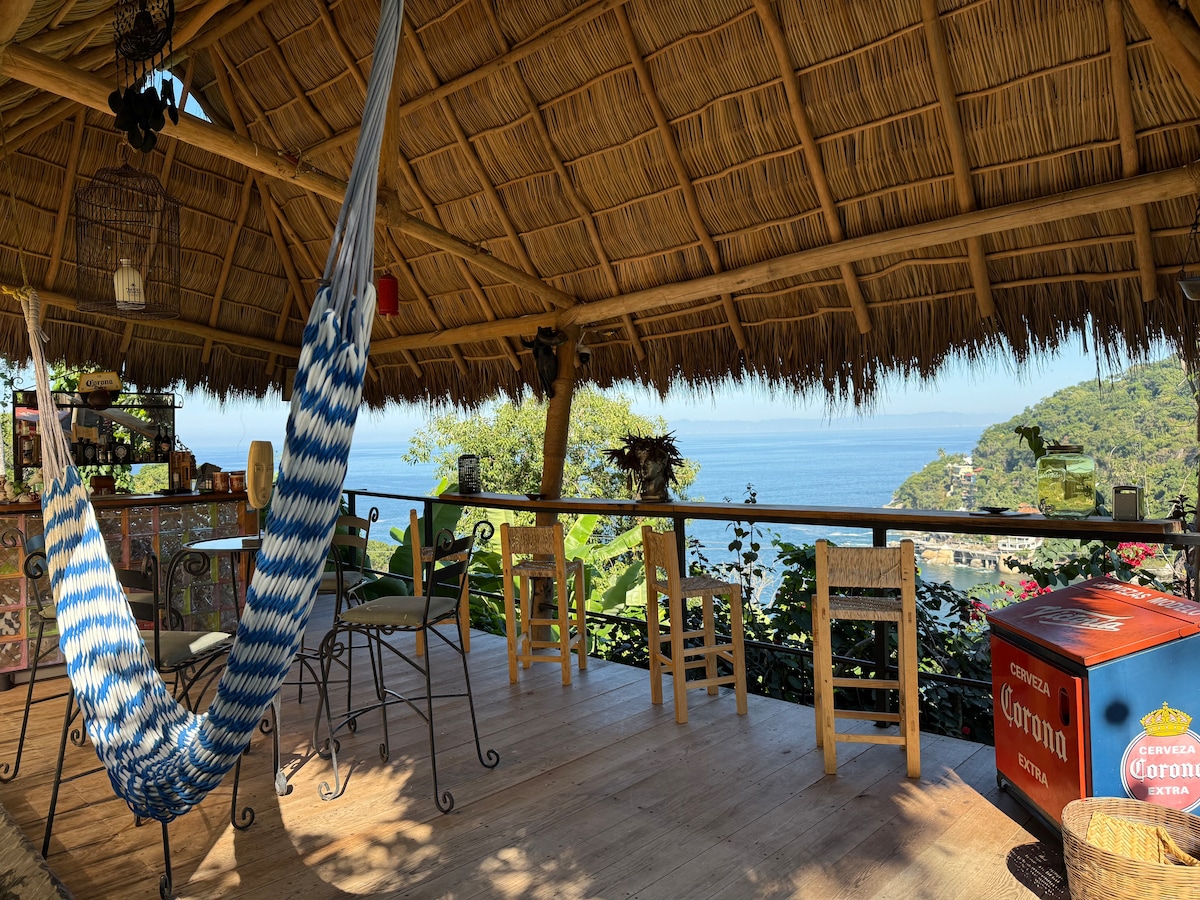 A spacious outdoor dining area is framed by a thatched roof, featuring a long bar with seating and wooden chairs. A blue and white hammock is gently swaying, with panoramic views of the ocean and lush greenery visible in the background.