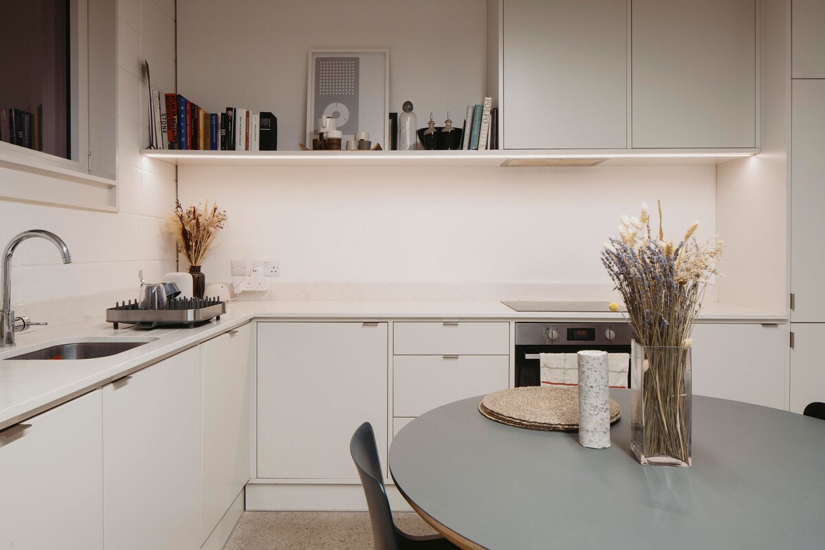 A modern kitchen space is featured, showcasing sleek white cabinetry and minimalist design. A round table with a neutral tone is set, accompanied by a vase of dried flowers and a small decorative item. A well-organized countertop holds dishware and kitchen appliances.