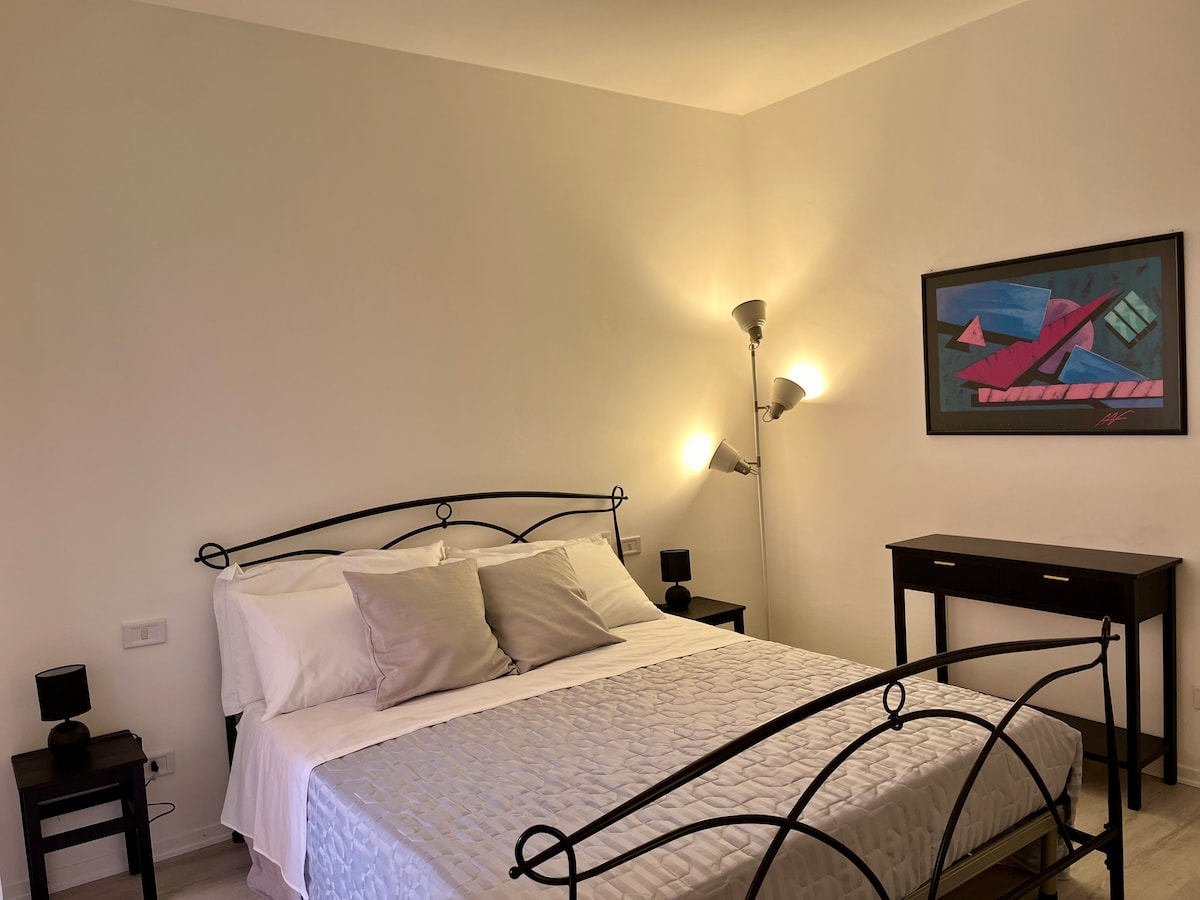 A double bedroom is featured, equipped with a spacious bed and two bedside lamps. A minimalist wooden dresser is positioned against the wall, while modern artwork adds a touch of color. Soft lighting from wall fixtures enhances the room's overall ambiance.