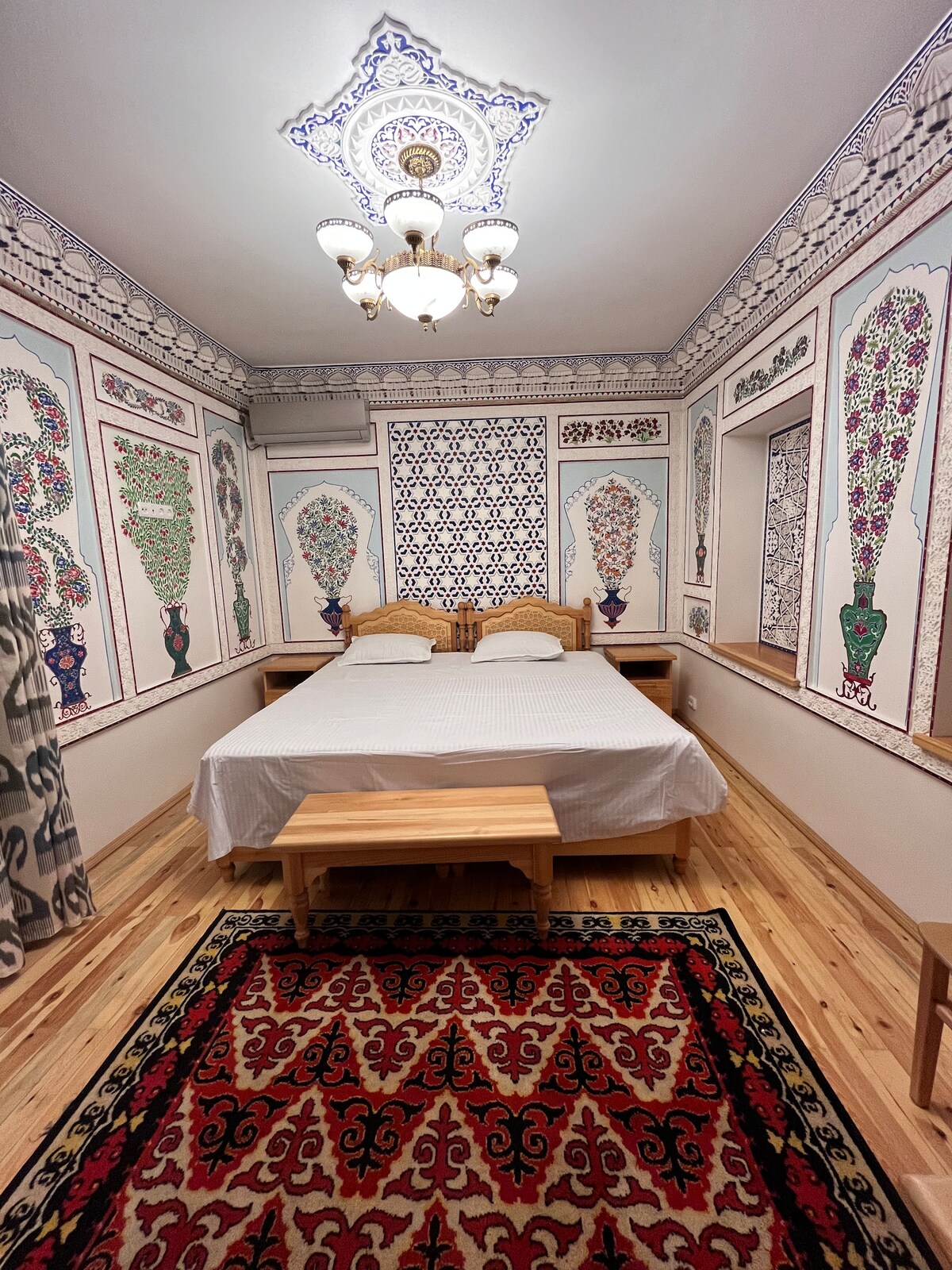 A cozy room features a spacious bed with linens neatly arranged. The walls are decorated with intricate eastern patterns in vibrant colors, complemented by a patterned area rug on warm wooden flooring. A stylish light fixture hangs from the ceiling, illuminating the space.