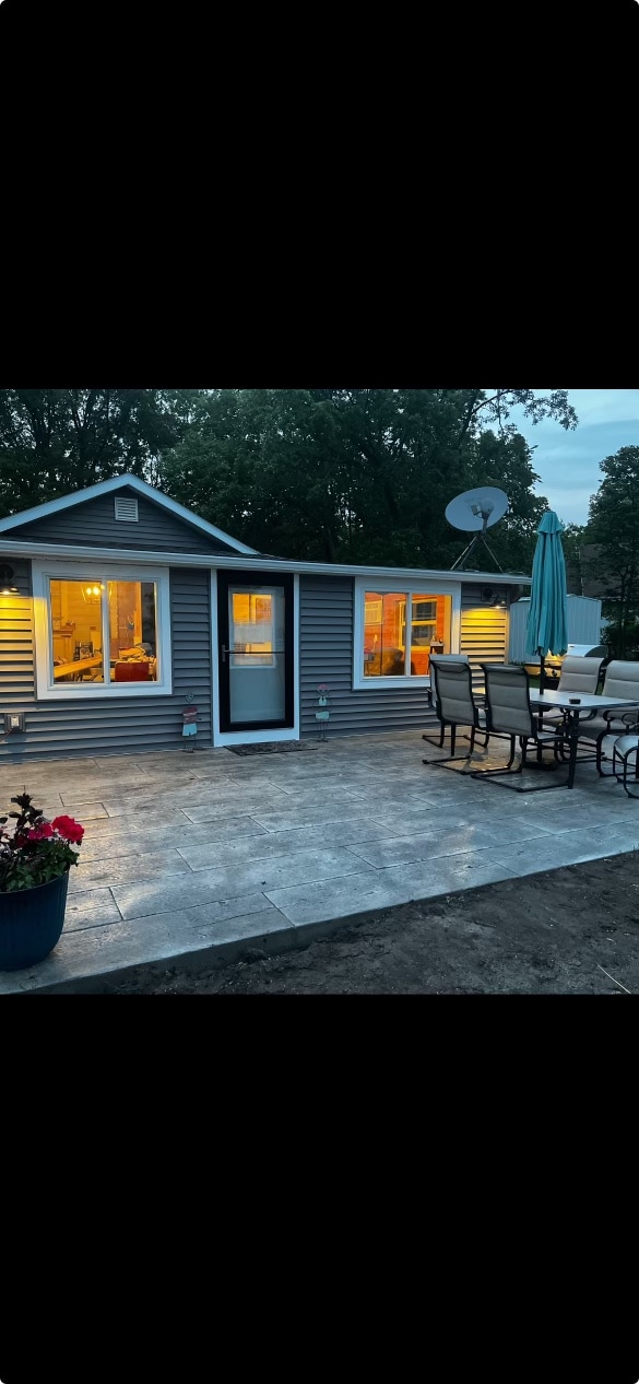 A welcoming cabin exterior is illuminated by warm light from the windows. The patio features a dining table with chairs and an umbrella, set on a well-maintained concrete surface. Flower pots and a satellite dish are visible, contributing to the inviting atmosphere.