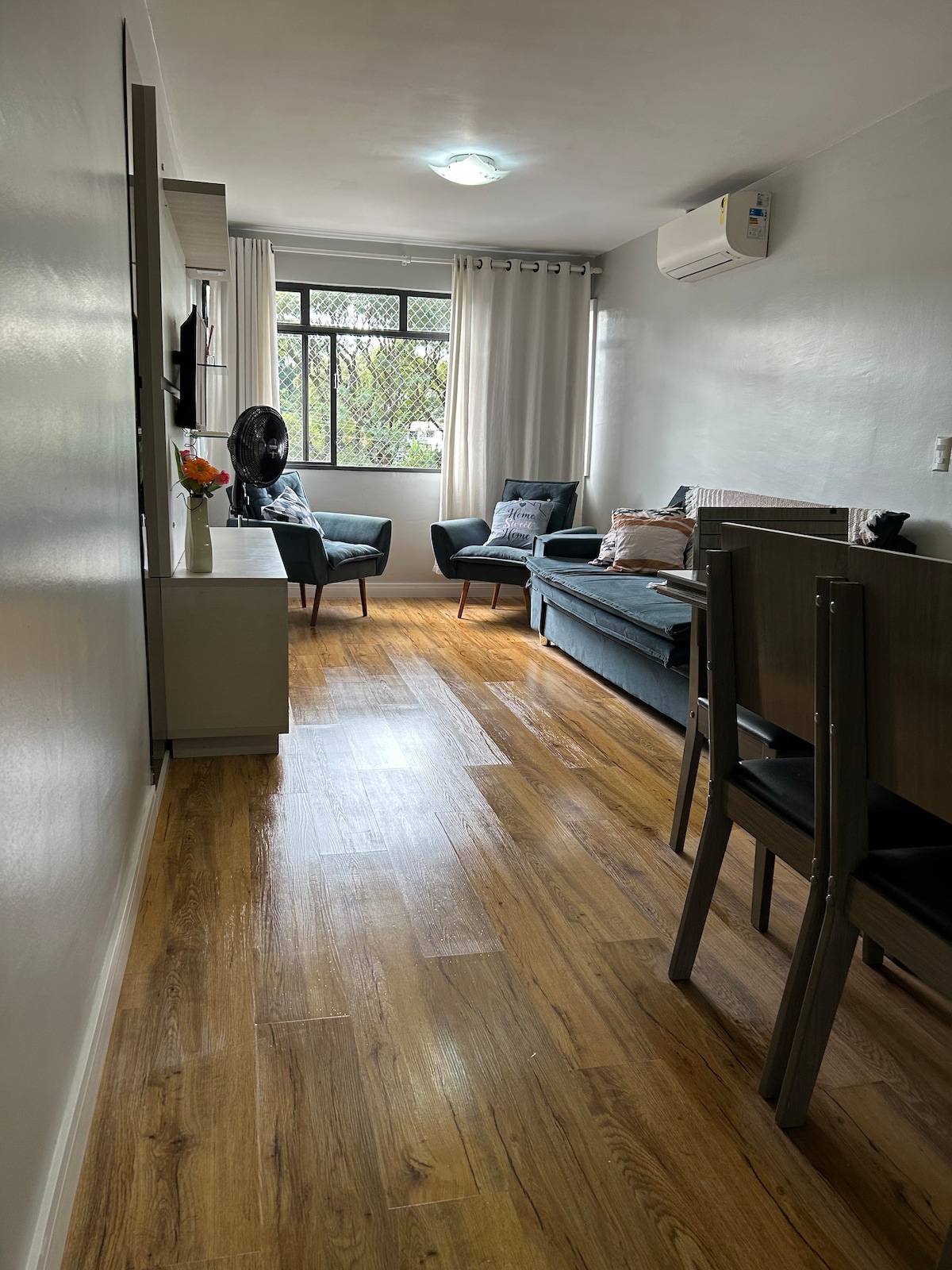 A narrow living area features a light wooden floor and neutral-colored walls. Two armchairs are positioned by a window, allowing natural light to filter through sheer curtains. A small dining table with chairs is visible, alongside a television mounted on the wall.