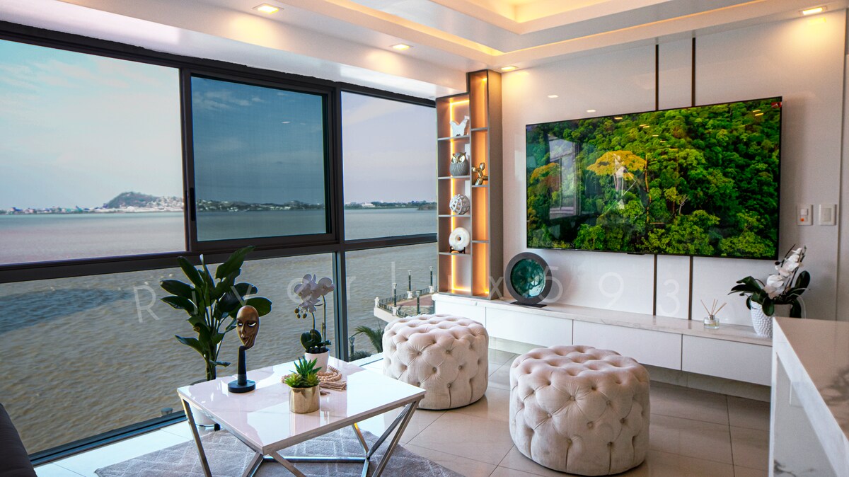 A modern living area features large windows providing a panoramic view of the Río Guayas. A stylish white coffee table is centered on the light-colored floor, accompanied by tufted ottomans. A flat-screen TV is mounted on the wall, showcasing a vibrant nature scene.